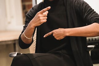 Lindsey (the article author) sits in a chair wearing a chic all-black outfit. Her index fingers point in opposite directions across her body as she wears a focused expression.