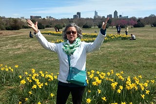 Photo of Mom in front of flowers in Dix Park in Raleigh with the skyline in the background. Her arms are raised to greet the world.