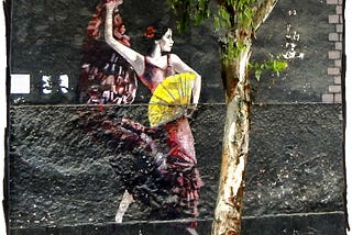 Photo of street art taken in Lima, Peru. It depicts a woman in a red dress dancing while holding a yellow fand. She’s on a gray wall facing a tree.