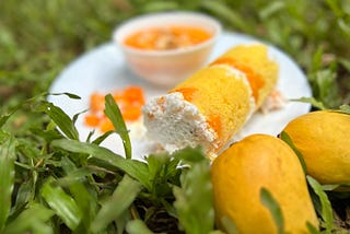 On a white-plate, a cylindrical rice cake with yellow color is served with visible mango cut in between them. Around it, a bowl of mango puree, cuts of mangoes, and two mangoes are placed.