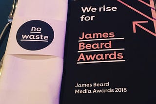 On the left, a linen napkin wrapped in a holder that says “no waste” on the right is a black printed program with pink writing that says “We rise for” James Beard Awards, James Beard Media Awards 2018.