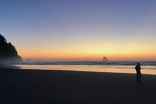 A sunset along the water. Rainbow faded sky and a glistening sandy shore, with the silhouette of a person in the corner.