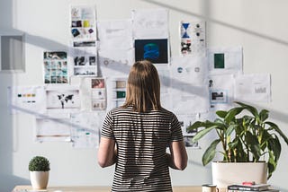 A person stands back to us, in front of a wall with design artifacts tacked up on it.