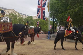 Three horses walking past, two of which are carrying silver kettle drums with their musician riders; one in front is carrying a soldier.