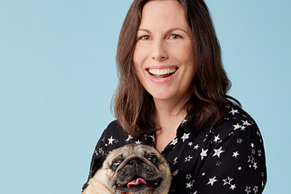 Image shows a lady with long dark hair, smiling and cuddling a pug dog