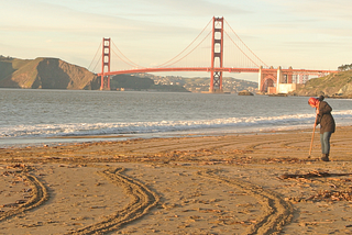 View of the Golden Gate Bridge from Baker Beach at sunset with woman raking sand in foreground
