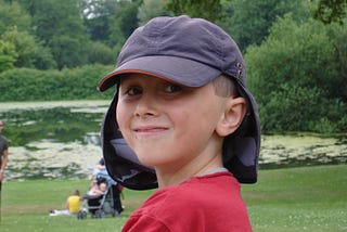 A young boy around 10 years old. He has a red t-shirt and a blue cap with a sun guard protecting his neck. He is looking at the camera and smiling. People are in the background admiring a lake, and the boy is surrounded by trees.
