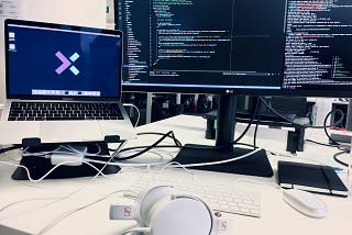 a white desk with a macintosh on an stand, an external monitor on its side, a white keyboard and a white headset over the desk