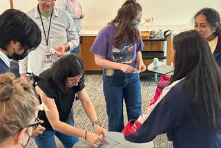 Eight people standing and looking down at a desk with a very small battery powered robot; smiles abound, and a snack cart in the background.