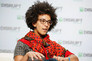 Timnit wearing a red patterned scarf and a grey sweater, speaking on stage.