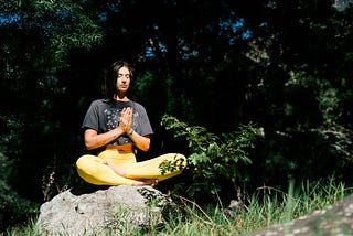 Dark-haired faired skinned woman sits criss-crossed on a rock with her hands clasp together in a yogic prayer medittive position outside, with trees as the background scenery.