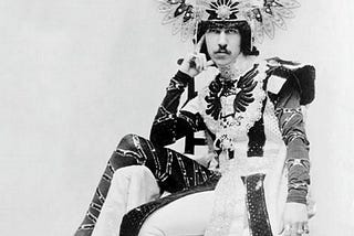 Paget c. 1900, photographed by John Wickens. He’s wearing a winged headdress, and an extraordinary, elaborate skirted costume and tights