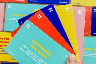 A thumb holds a selection of brightly coloured PR prompt cards in green, red, blue, yellow and pink on top of a backdrop of cards.