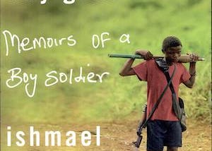 This is an image of the cover of the book “A Long Way Gone: Memoirs of a Boy Soldier” by Isnmael Beah. The image shows a young boy wearing torn shoes and dirty clothing carying a weapon over his shoulders. He is walking alone down a dirt path by a grassy hill.