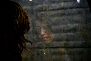 A sad woman looks out of a window, her reflection captured in the glass.