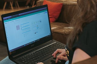 Uma mulher sentada em um sofá, digitando em um notebook ligado em uma página do Miro.
