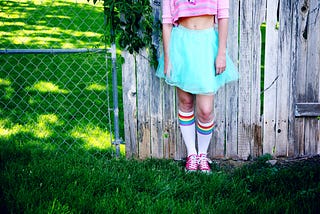 On an outside lawn, leaning against a fence which is 1/2 chain link (left side) and 1/2 wooden boards (right side) leans a caucasian person. Torso to feet are visible, head is not. Pink half shirt, bare midriff, turquoise poofy skirt, and rainbow kneesocks leading to pink converse sneakers are worn.