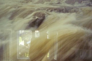 In a photograph of a river cascading over rocks at dawn, a reflection of a figure stands before a window.