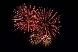 Four firework shells exploding in the sky in dandelion fluff shape. The two largest are pink and gold, the two smaller ones just below are gold.