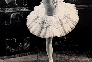 A black and white photo of a ballerina standing a top a metal bucket stand, in a lace tutu. The black tiles in the background reflect the white of the ballerinas tutu
