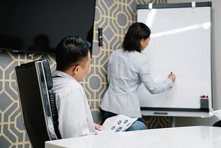A man looking on while sitting while a woman is standing at a whiteboard mocking something up.