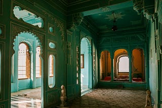 A view into several rooms of a beautiful Moroccan space, with sea-foam/teal walls, high contrast tile flooring, ornately arched doorways, and sunlight pouring in through windows.