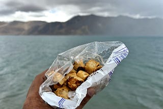 An opened pack of savory snacks with a backdrop of Pangong Lake and the Himalayas