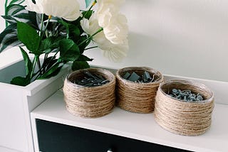 A white desk with twine wrapped plastic containers that were repurposed and to use for holding stationery items such as paper clips, push pins, and binder clips. There is also a little bouquet with white flowers and green leaves.