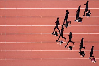 Aerial photograph of runners mid race on an athletics track, with silhouttes cast by the sun, running in ‘V’ formation with the lead runner in centre.
