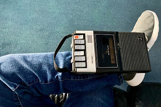 A cassette recorder balanced on someone’s leg. The person is wearing blue jeans and grey trainers.