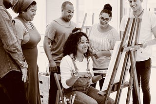 Woman of color sitting at an easel with a crowd appreciating her work. Envato Elements Item ID: EPMAC2S. License available upon request.