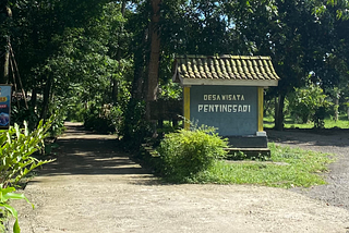 Salah satu prasasti yang memberikan keterangan “Desa Wisata Pentingsari” terletak di bumi perkemahan milik dusun. Buper ini berdekatan dengan sekretariat desa wisata dan warung makan Omahe Simbhok. Suasana rindang nan teduh jadi pengalaman wisata yang mudah ditemukan kala melawat ke sana. Tampak lanskap sebuah prasasti berwarna abu-abu dengan tulisan putih “DESA WISATA PENTINGSARI” berikut rerumputan di sekitarnya. Sumber gambar: Haidar Karel