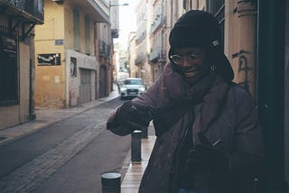 Toyin during their trip. They are wearing a coat and hat, holding a cig and grinning on a tight street in Perpignan, France