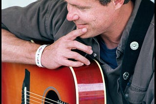 Photograph of singer-songwriter Dave Carter resting on his guitar, which is resting on his knee, as he listens to another songwriter perform a song at a workshop stage at the Falcon Ridge Folk Festival on 29 July 2000, two years before he died. Photo copyright 2023 by Patrick T. Power. All rights reserved.