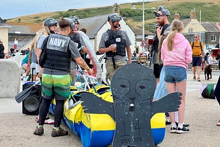 A raft with a great white head at the back and a seal at the front rowed by Navy personnel with seal mask/hats on
