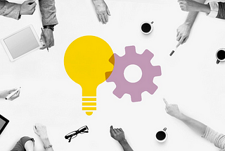 This picture conveys about a group of designers working together on a table and on that table there is an icon of a bulb(yellow) and a gear(light purple).