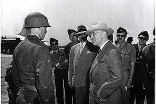 President Harry Truman stands in front of a group of soldiers on an airplane tarmac. He wears a brown suit, a white hat, and has his hands in his suit pockets. His head is turned to look at a soldier in a uniform and helmet, whose face we see in profile.