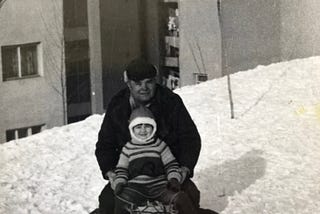 Black-and-white photo of young child with older man on sled, apartment buildings behind them