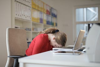 A woman with her forehead on her desk in front of her.