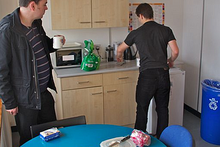 Two men in an office kitchen making coffee and tea