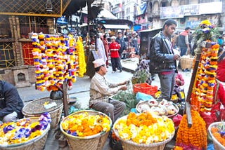 A Nepalese man sits on a stool with garlands of marigolds and chrysanthemums hanging on a rack beside him. There are also baskets full of colorful flowers in front of him. A man is looking at another rack of garlands on the other side of the seller.