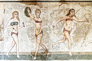 Mosaic portraying three young girls wearing what look like bikinis: one is holding small dumbbells, one is in the act of throwing a discus, the other is running. Two of the girls have blonde hair, the third has brown hair. The discus throwing girl is wearing golden bracelets, necklace and ankle bracelets