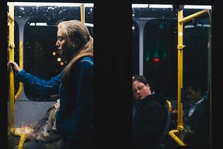 A woman standing alone on a bus at night, holding onto the yellow metal pole in front of her and looking down