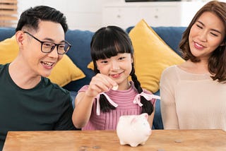 Two parents smiling with a child who is placing a coin in a piggybank.