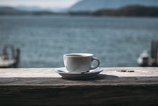 A white espresso cup sits on weathered wood in front of a beautiful ocean scene.