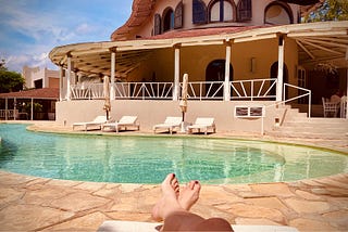 Image shows a pair of feet, resting on a lounger in front of a kidney-shaped pool, four empty loungers and a deck across the way.