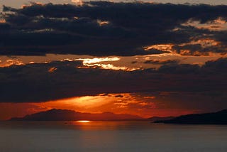 Final moments of shaft of sunset on water with distant islands. bands of dark clouds are fringed with silver. The sky closer to the water is a amalgam of fire and gold.