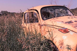 A yellow car in an abandoned field. The passenger side door is slightly open and the windshield is partially broken open.