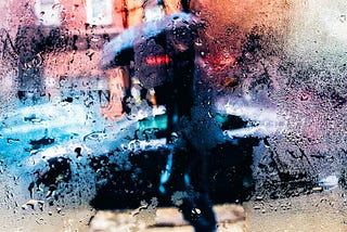 A window obscured by rain, and through it, there is a person dressed in black carrying a black umbrella.