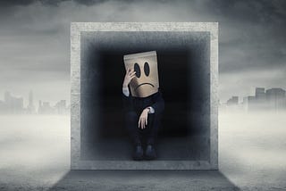 A man sat in a concrete box with a paper bag over his head displaying a drawn sad face.